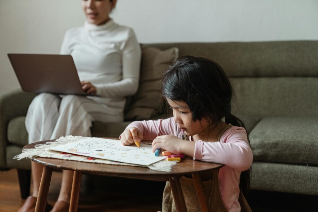 A mother works on a laptop as her young daughter colors at a coffee table.