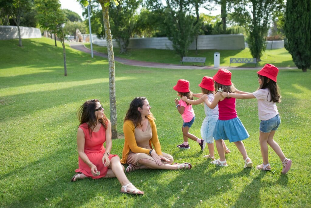 Happy children playing in a sunny park with two mothers sitting nearby, enjoying the outdoors.