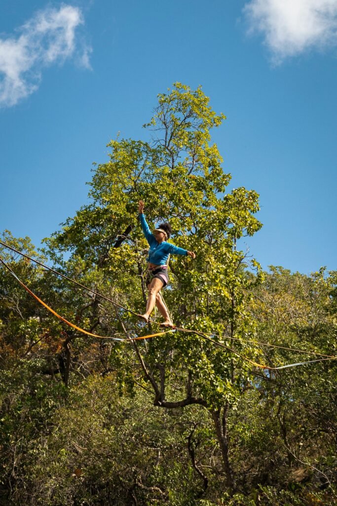 A woman in a blue shirt balances on a tightrope in a sunny forest park, showcasing outdoor adventure and agility.