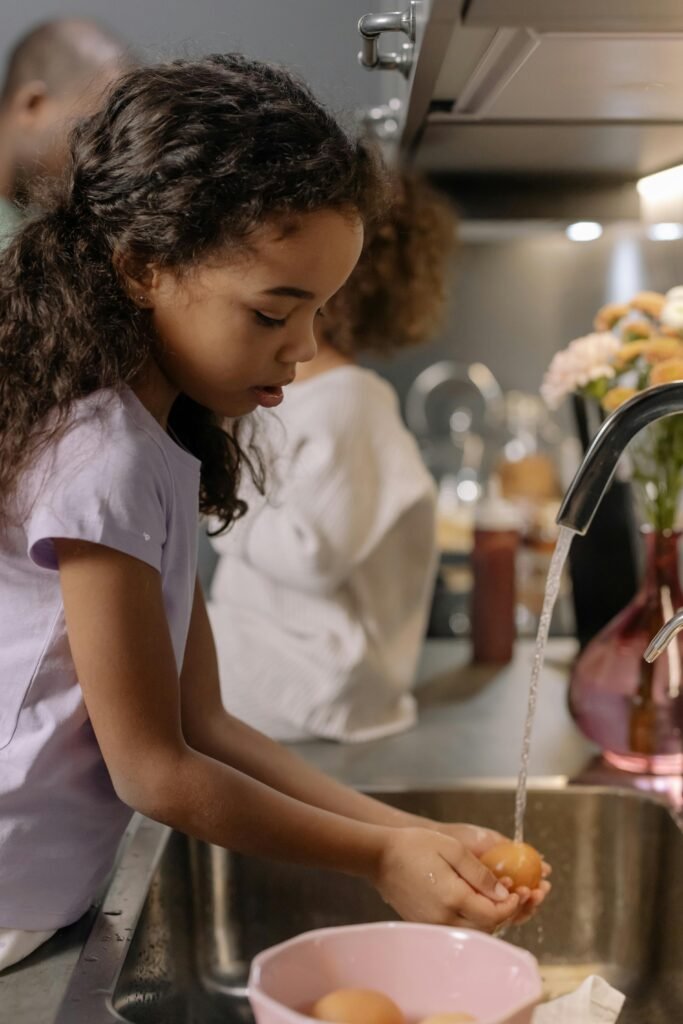 A young girl with curly hair washes eggs under running water in a modern kitchen.